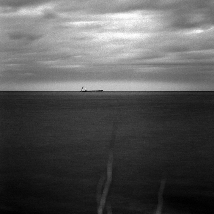 A cargo ship is visible on the horizon of a calm sea under a cloudy sky.