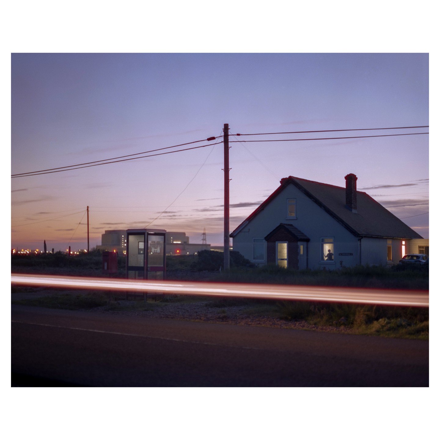 A house with illuminated windows stands beside a road with light trails at dusk, with a telephone booth and industrial buildings in the background.