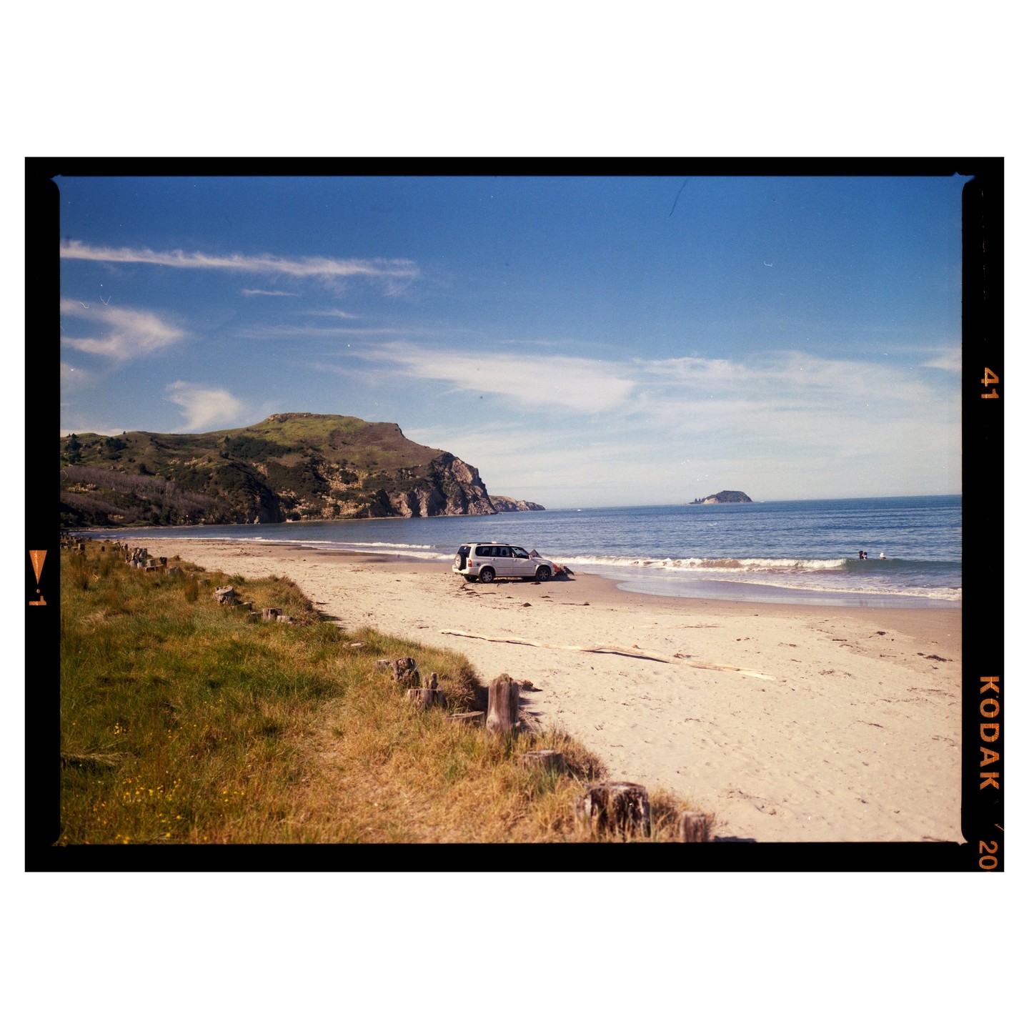 A white vehicle is parked on a sandy beach next to the ocean, with a large hill in the background and a small island in the distance.