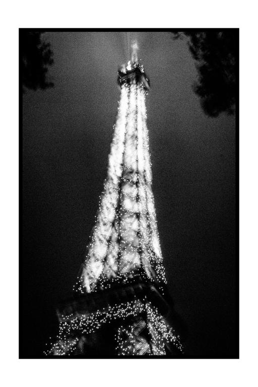 The Eiffel Tower is illuminated at night, with trees framing the top corners of the image.