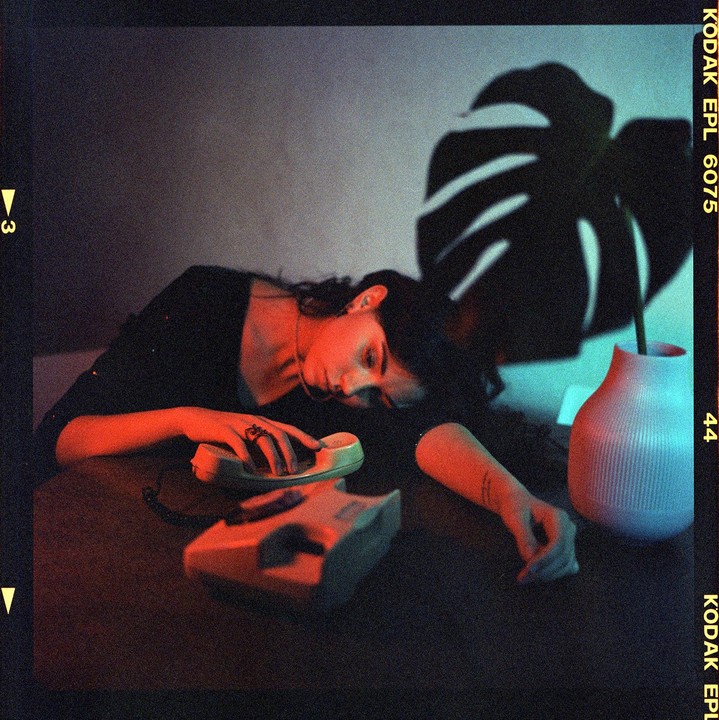 A woman rests her head on a table next to a telephone, illuminated by red and blue light.