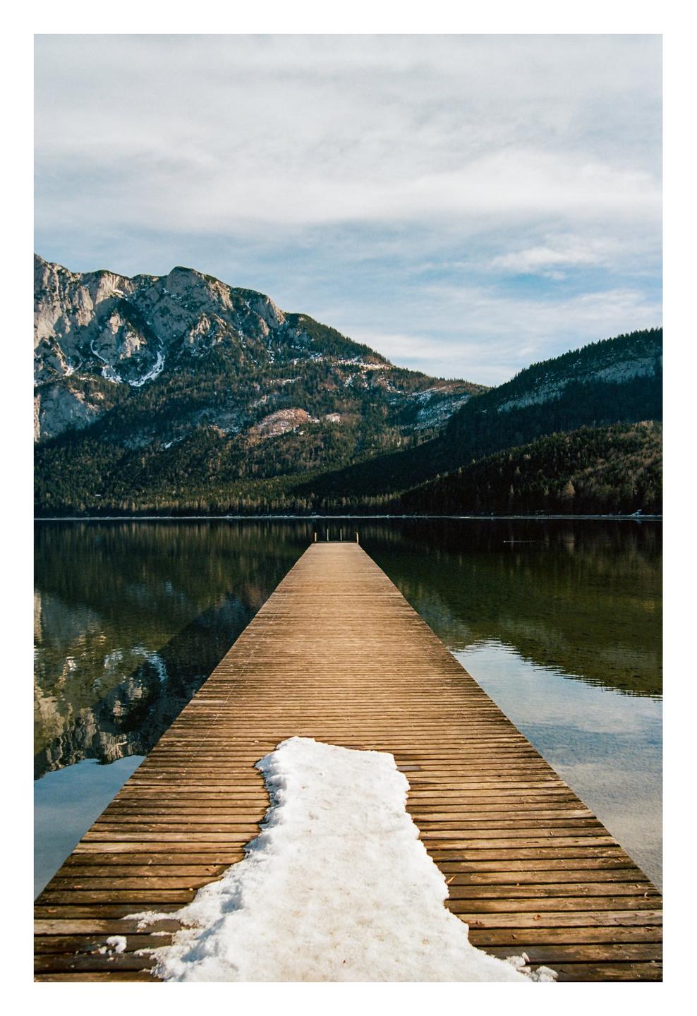 A wooden pier with a patch of snow extends into a lake surrounded by mountains and forest.