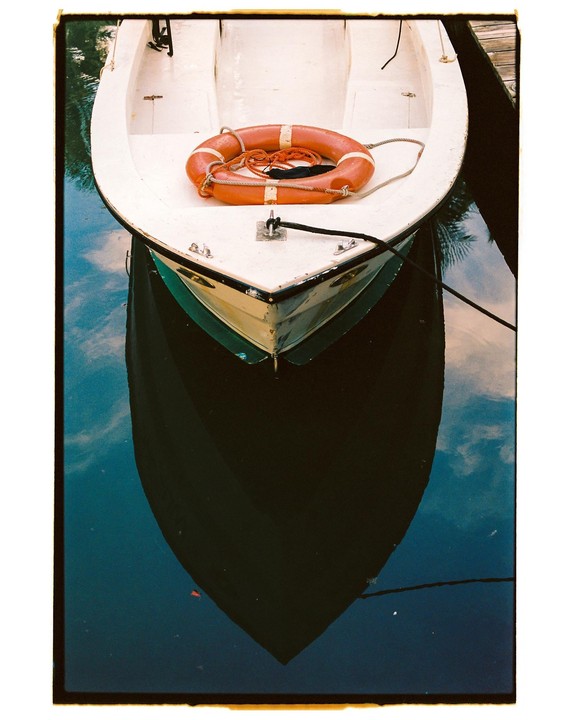 A white boat with an orange life preserver floats on dark water, reflecting the sky.