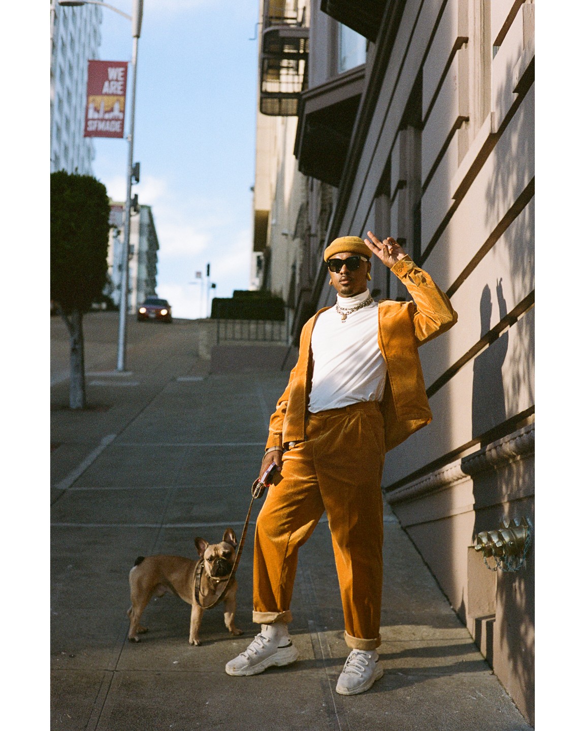 A man in a mustard suit and a french bulldog stand on a city sidewalk.