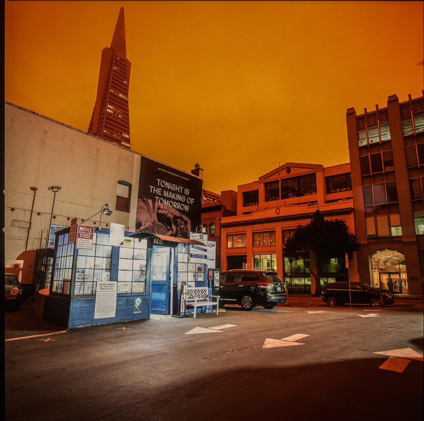 A tall, pointed skyscraper rises above a city street with buildings, parked cars, and a small kiosk under an orange sky.
