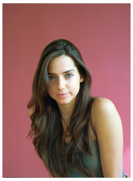 A woman with long brown hair and blue eyes looks directly at the viewer against a pink background.