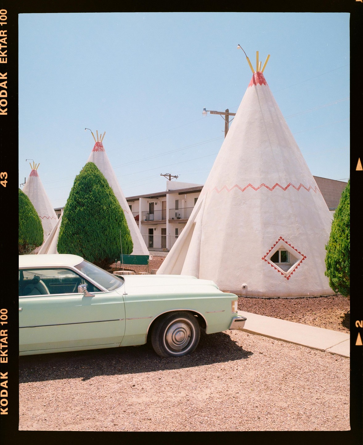 A light green car is parked in front of several teepee-shaped buildings with a motel in the background.