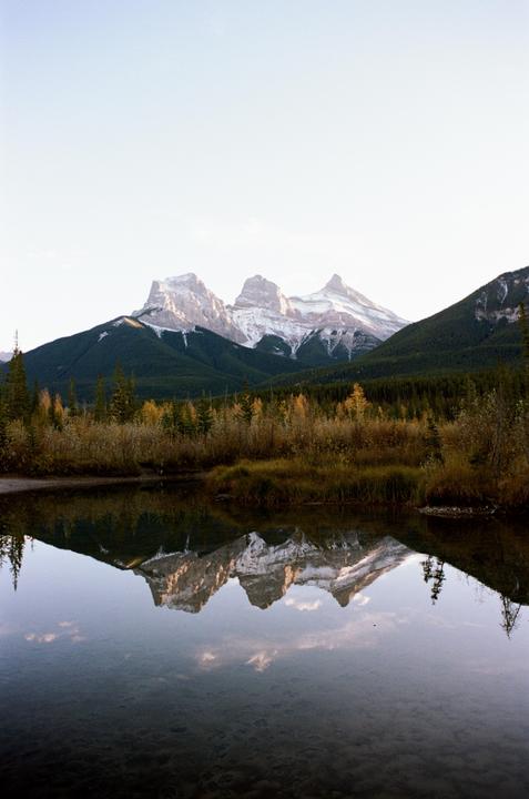 Snow-capped mountains are reflected in a calm body of water, surrounded by autumn foliage and evergreen trees.