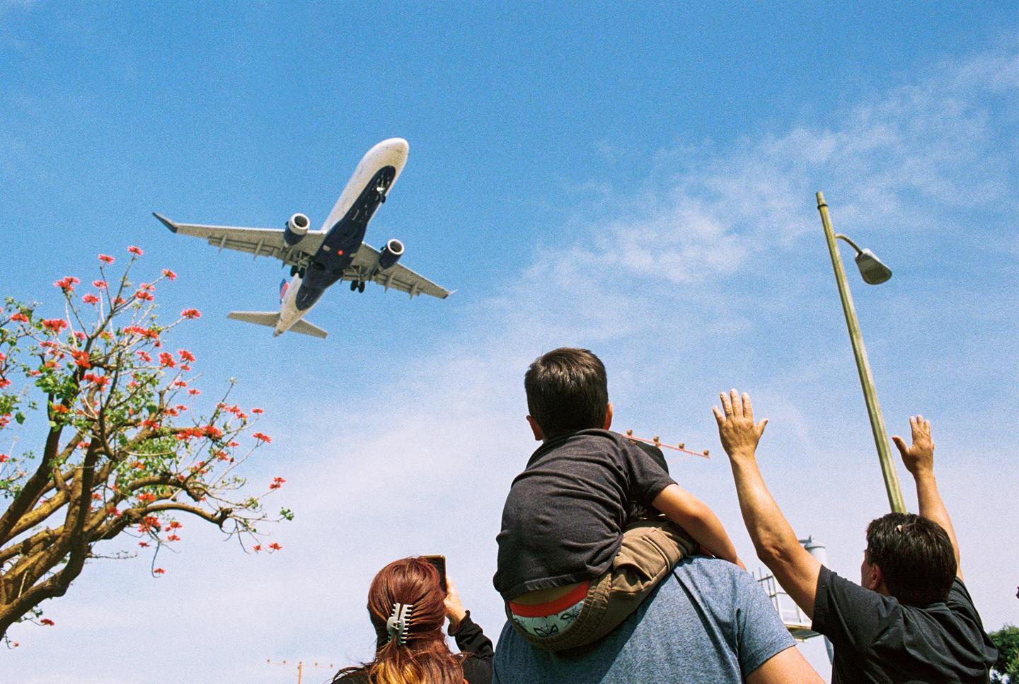 A family watches an airplane fly overhead, with a child on a man's shoulders and a woman taking a photo.