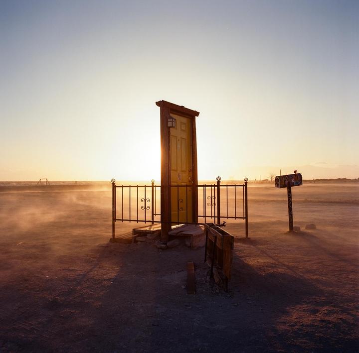 A yellow door stands alone in a dusty, barren landscape with a mailbox nearby during golden hour.