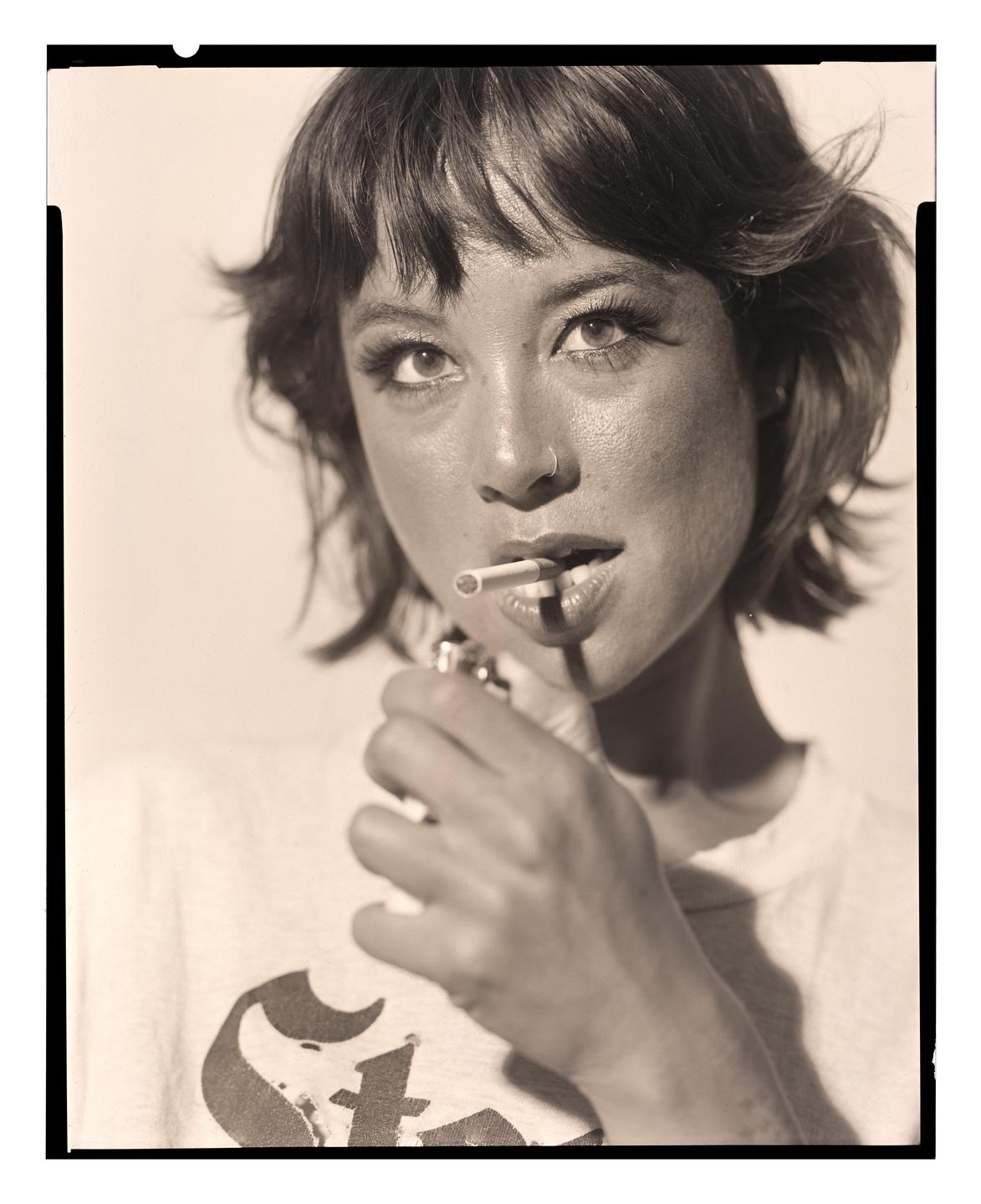 A woman with short hair holds a cigarette in her mouth and a lighter in her hand.