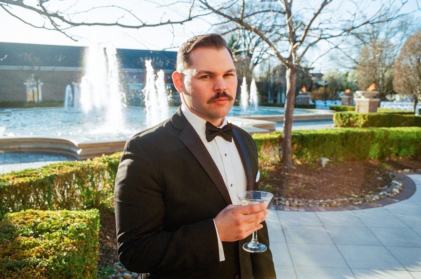 A man in a tuxedo holds a martini glass while standing in a garden with fountains and topiary.