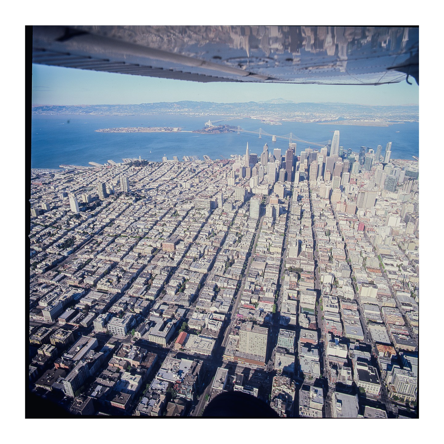 An aerial view of San Francisco shows the city grid, skyscrapers, and the bay with bridges and Alcatraz Island under a clear sky.