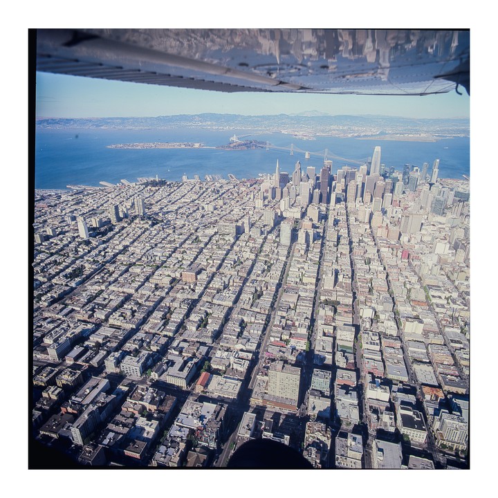 An aerial view of San Francisco shows the city grid, skyscrapers, and the bay with bridges and Alcatraz Island under a clear sky.
