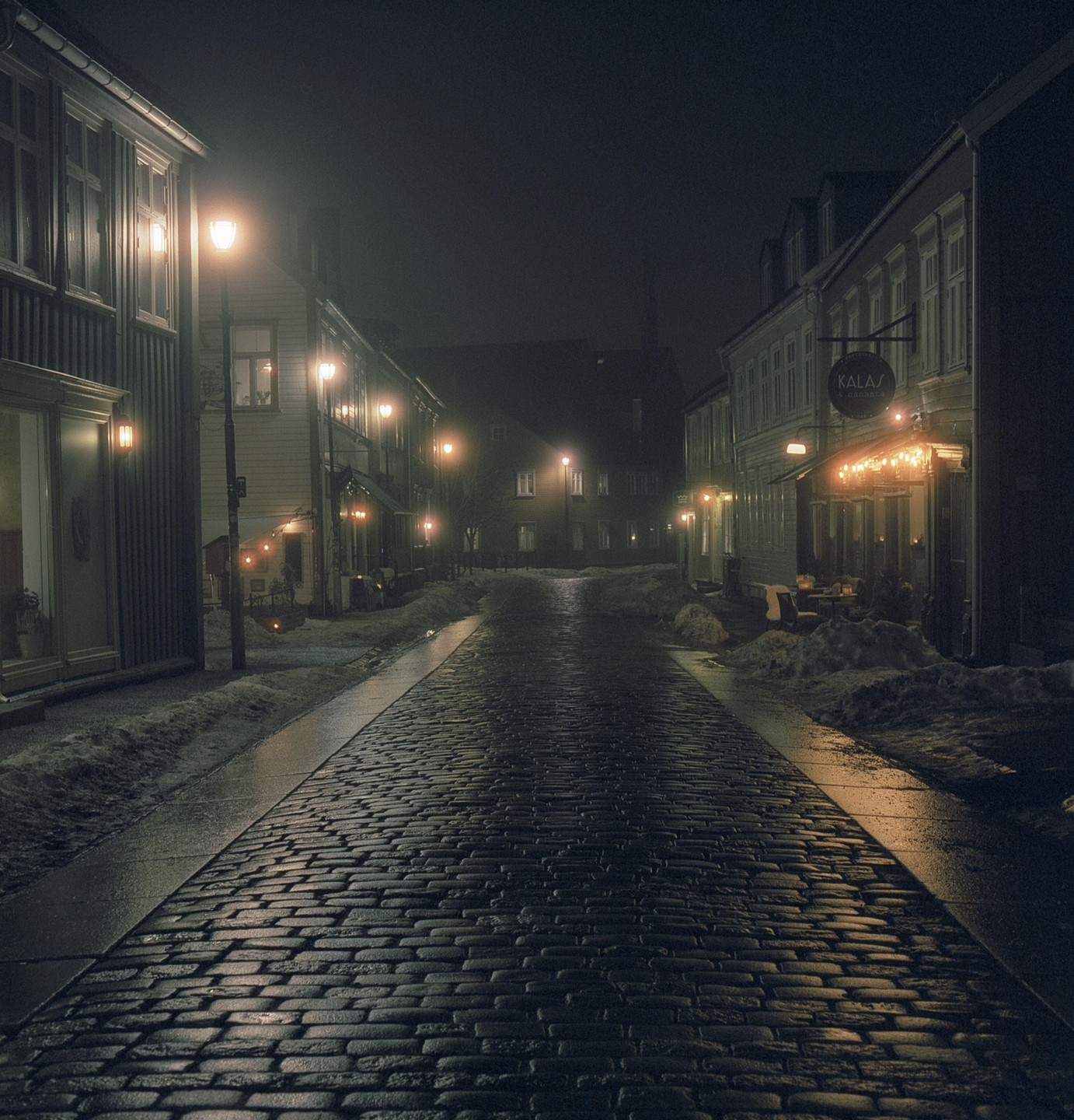 A cobblestone street is lined with buildings and streetlights at night, with snow piled on the sides of the road.