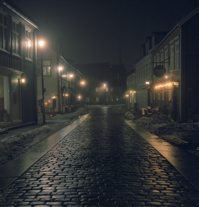 A cobblestone street is lined with buildings and streetlights at night, with snow piled on the sides of the road.