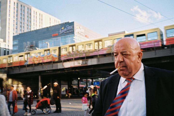A man with a cigarette in his mouth walks past a train on an elevated track in a city.