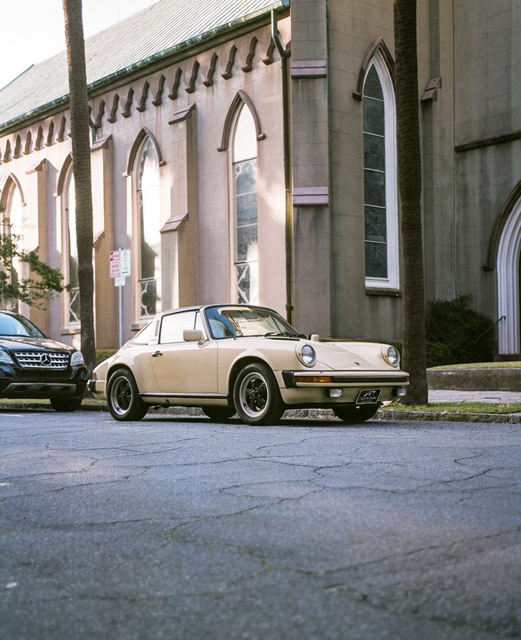 A beige Porsche is parked on a street in front of a church with tall arched windows and palm trees.