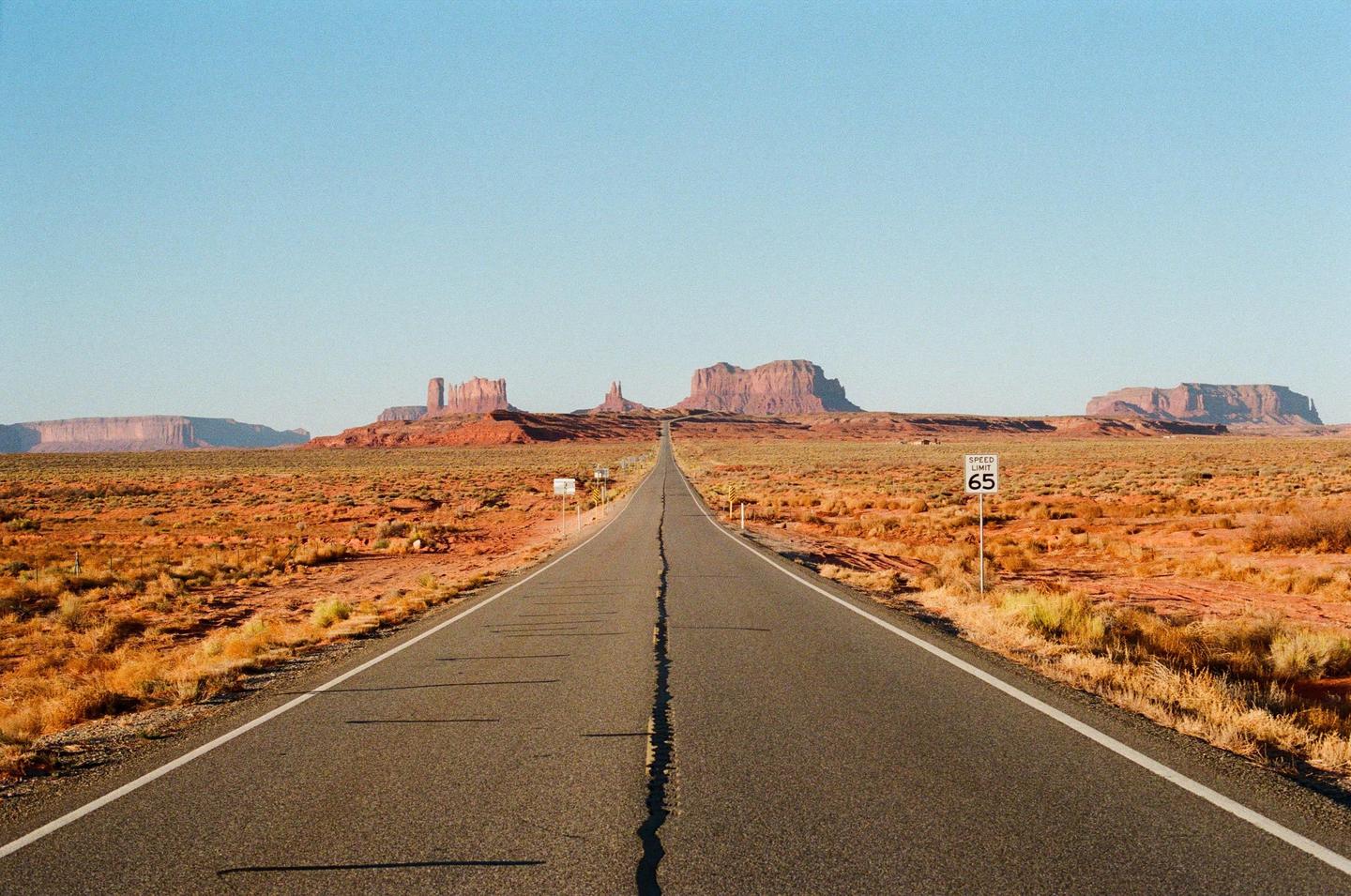A long, straight road stretches into the distance towards a series of large rock formations under a clear sky.