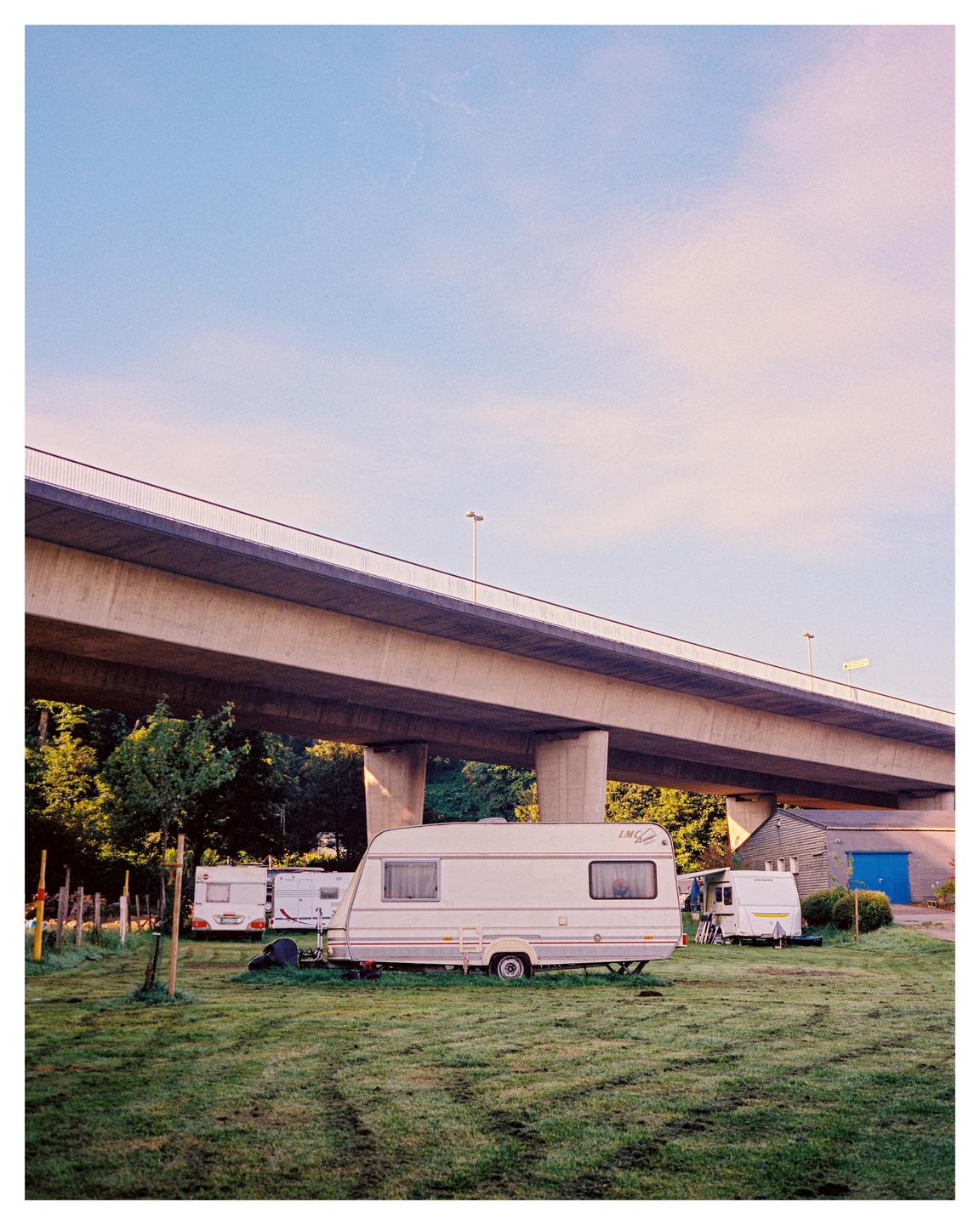 Several caravans are parked on a grassy field under a large concrete overpass.