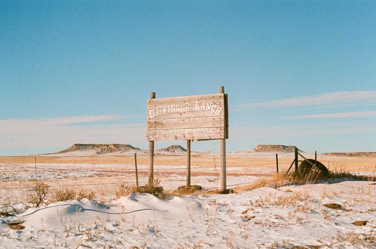 A wooden sign for Grisinger Ranch stands in a snowy field with distant mesas under a clear sky.
