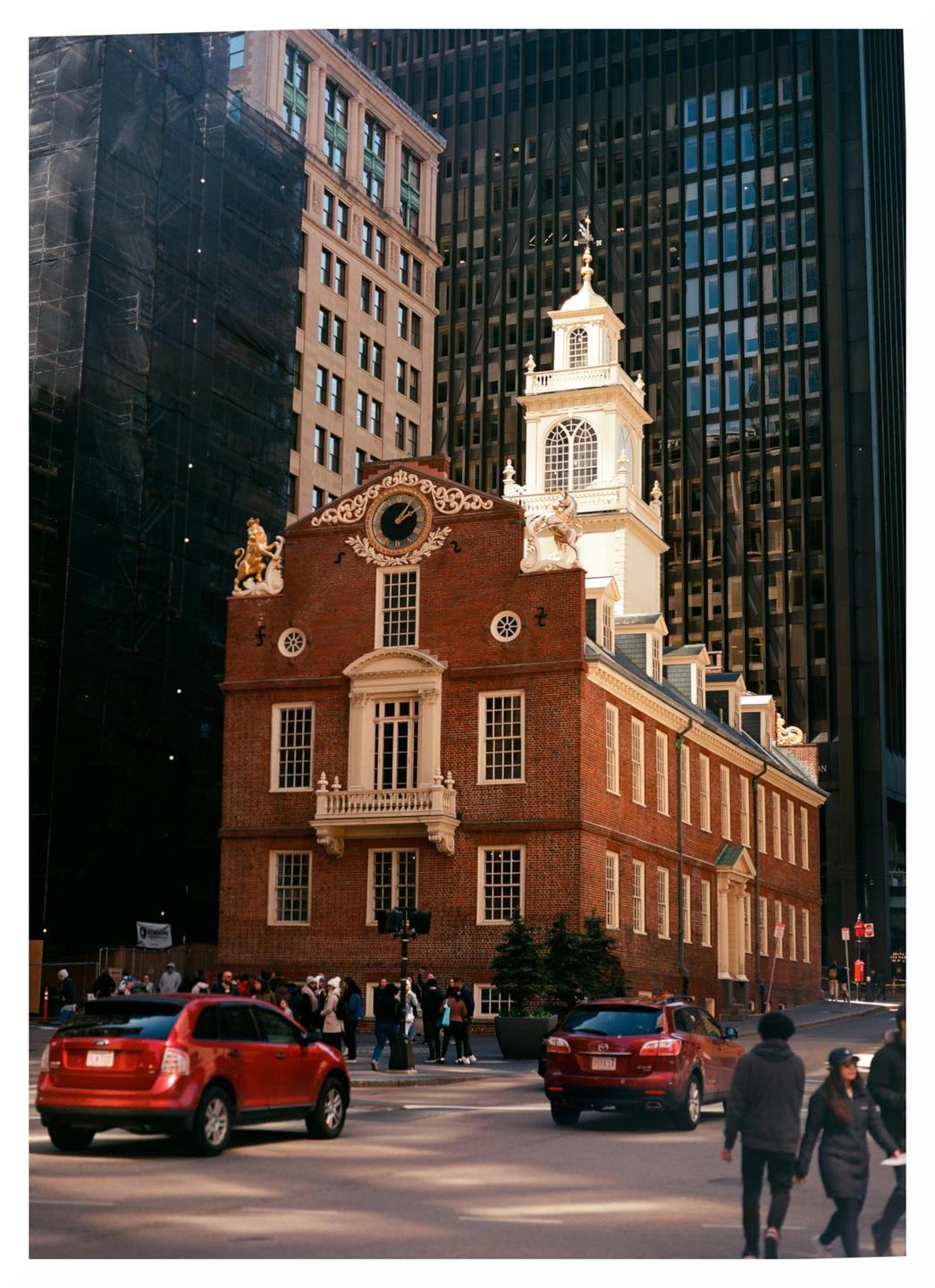A red brick building with a white cupola and clock tower stands among modern skyscrapers, with cars and people on the street below.