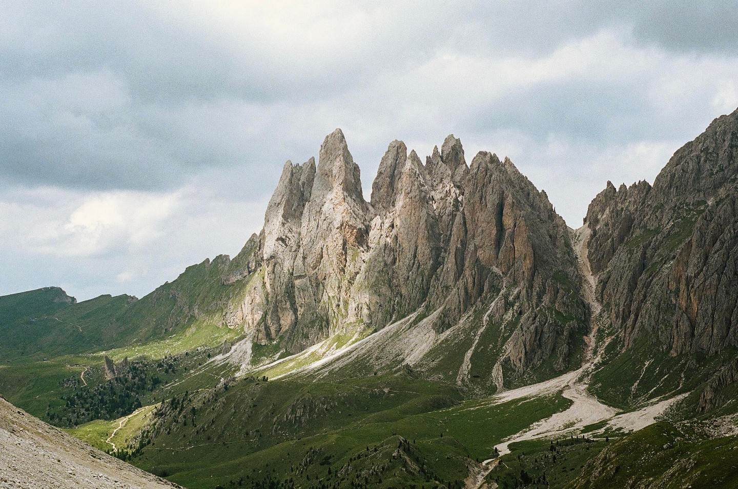 A dramatic mountain range with jagged peaks rises above a green valley under a cloudy sky.
