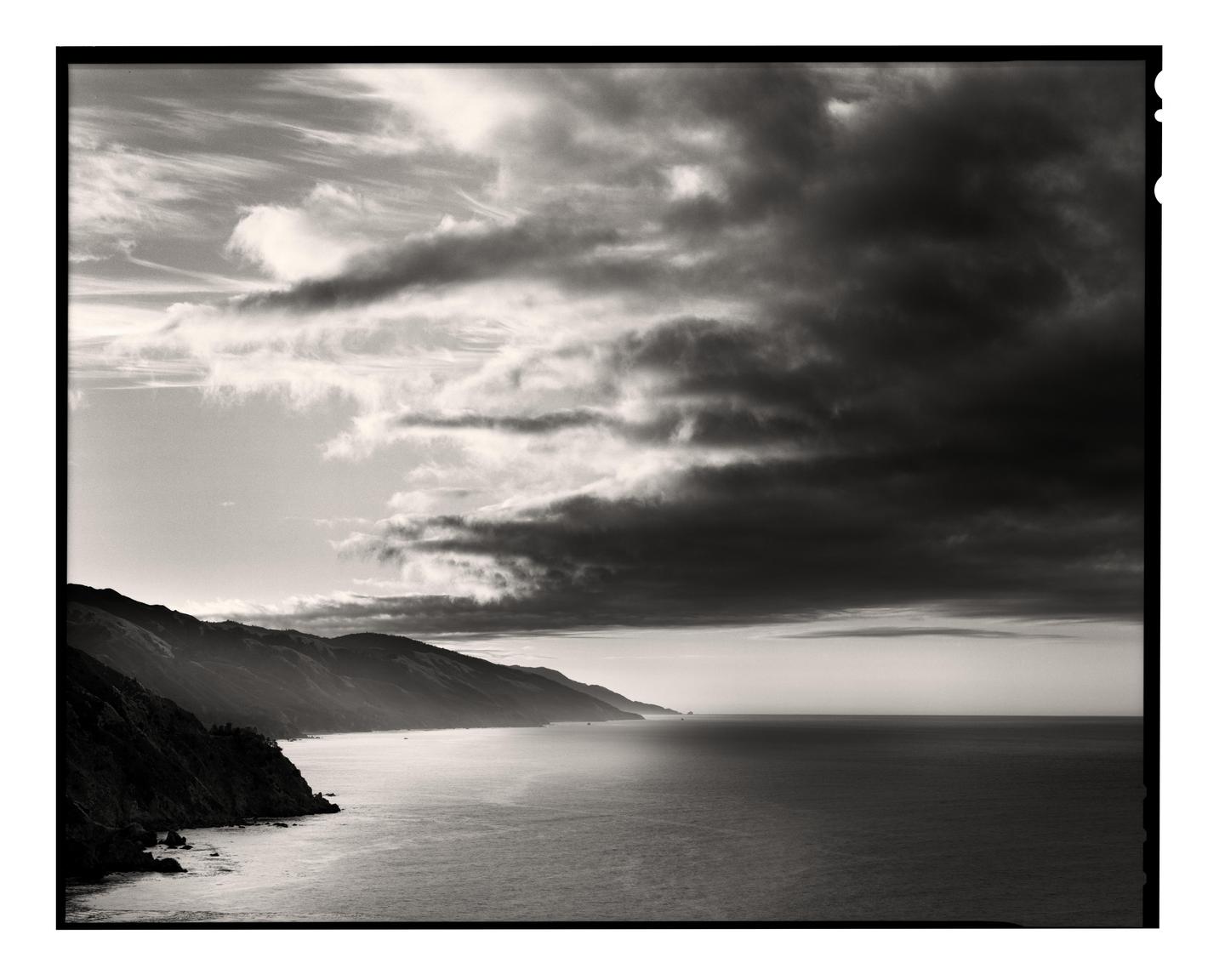 A dramatic black and white landscape shows a rugged coastline with mountains meeting the ocean under a sky filled with dark clouds.