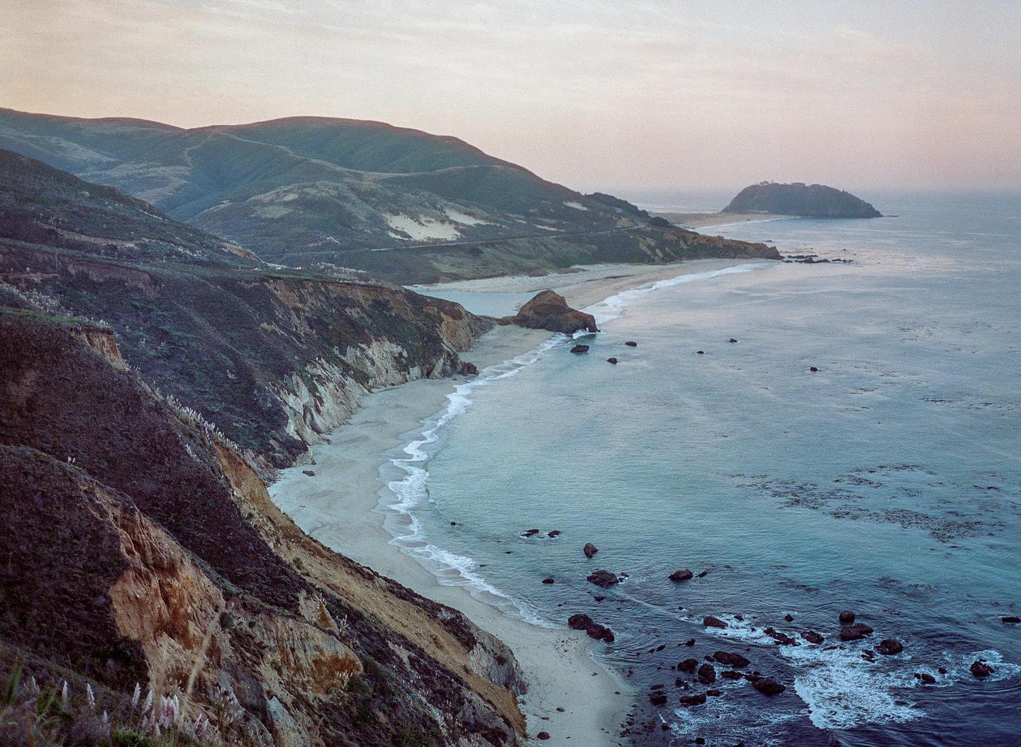 A long stretch of beach and ocean is visible from a high vantage point, with mountains in the background.