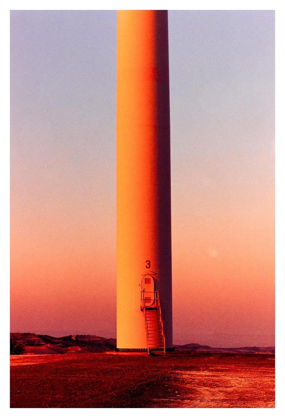 A wind turbine stands in a field at sunset, with a ladder leading to a door on its base.
