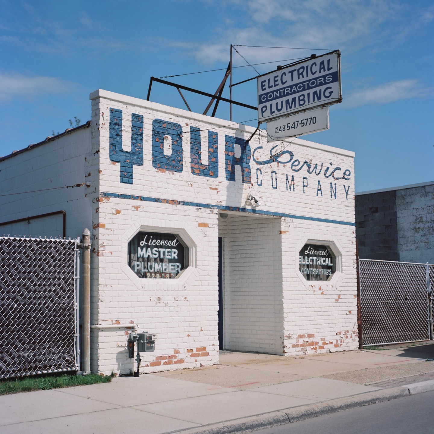 A white brick building with blue lettering for "Your Service Company" stands next to a chain-link fence under a clear sky.