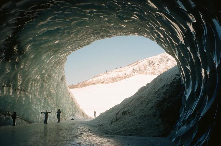 Three people stand inside an ice cave looking out at a snow-covered landscape.