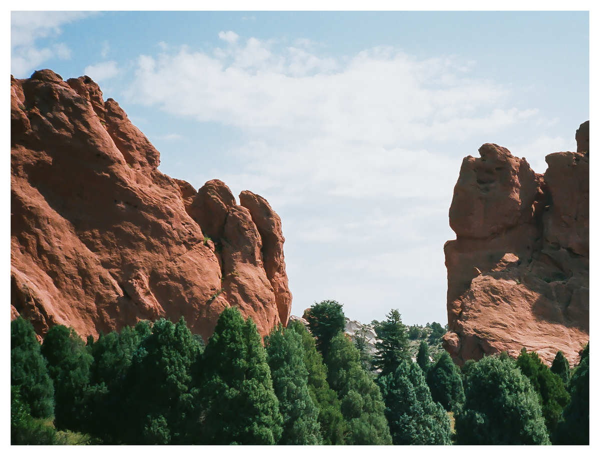 Two large red rock formations frame a gap filled with green trees under a cloudy sky.