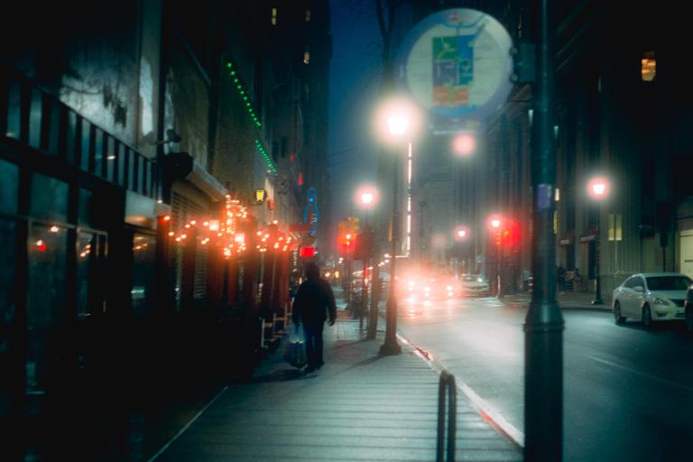 A person walks along a city sidewalk at night with cars driving on the street and lights illuminating the scene.