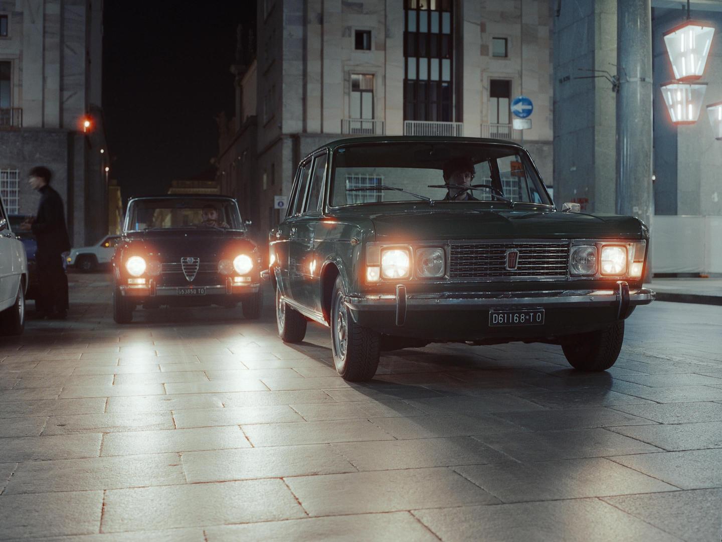 Two vintage cars with their headlights on are parked on a paved street at night, with a person standing next to one of the cars.