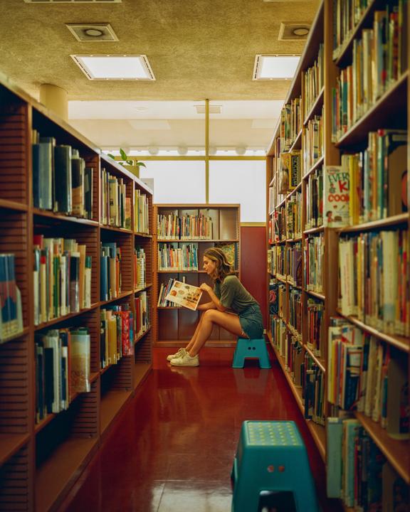 A woman sits on a stool in a library aisle, reading a book.