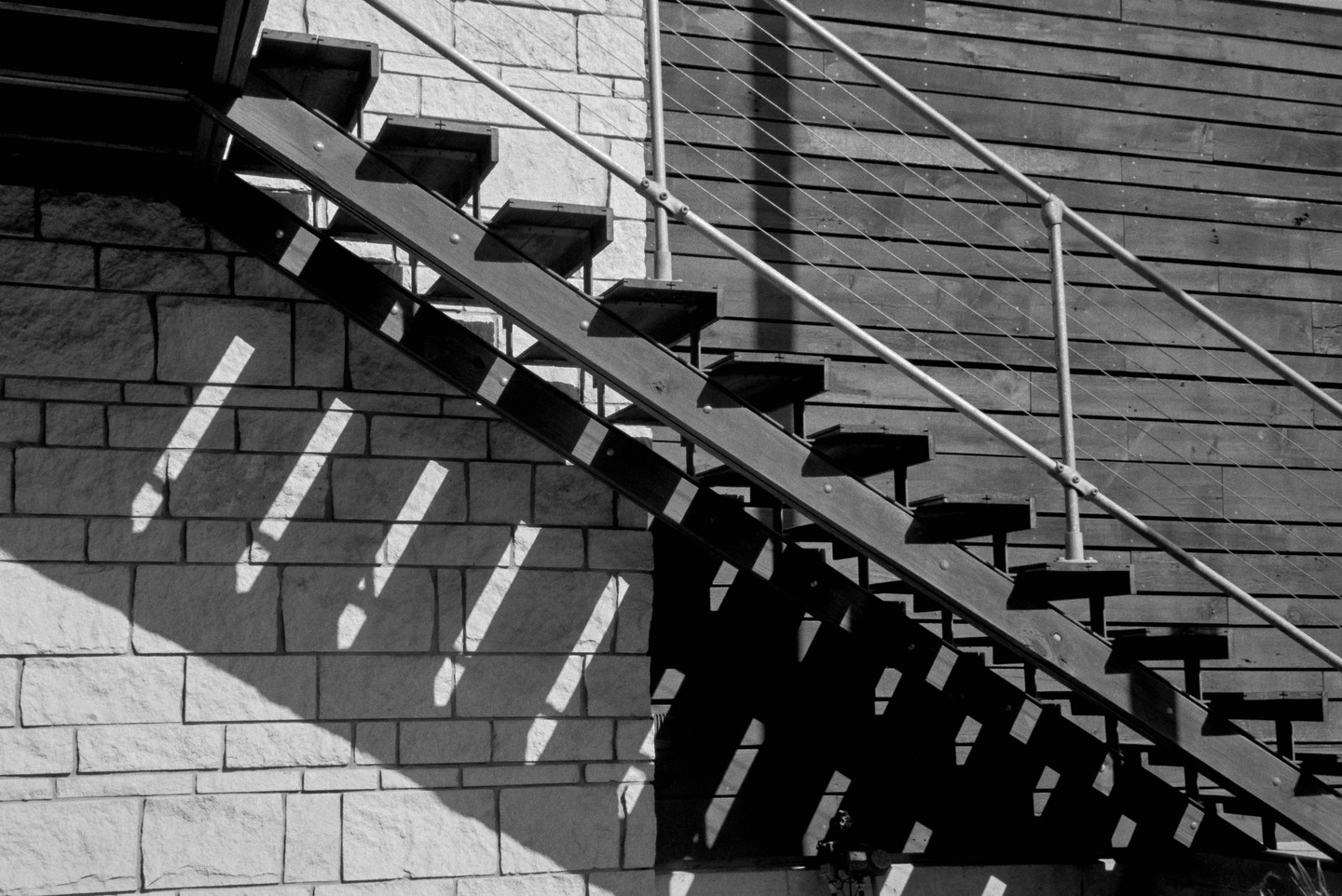 A wooden staircase with metal railings casts strong shadows on a stone wall and a wooden wall.