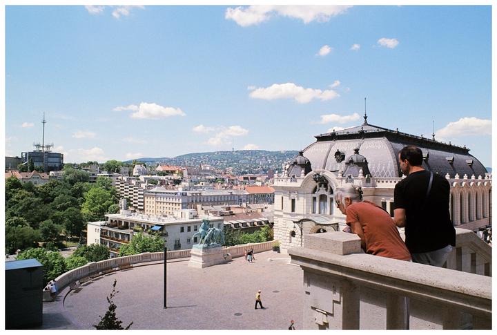 A man and a woman look out over a city from a balcony on a sunny day.