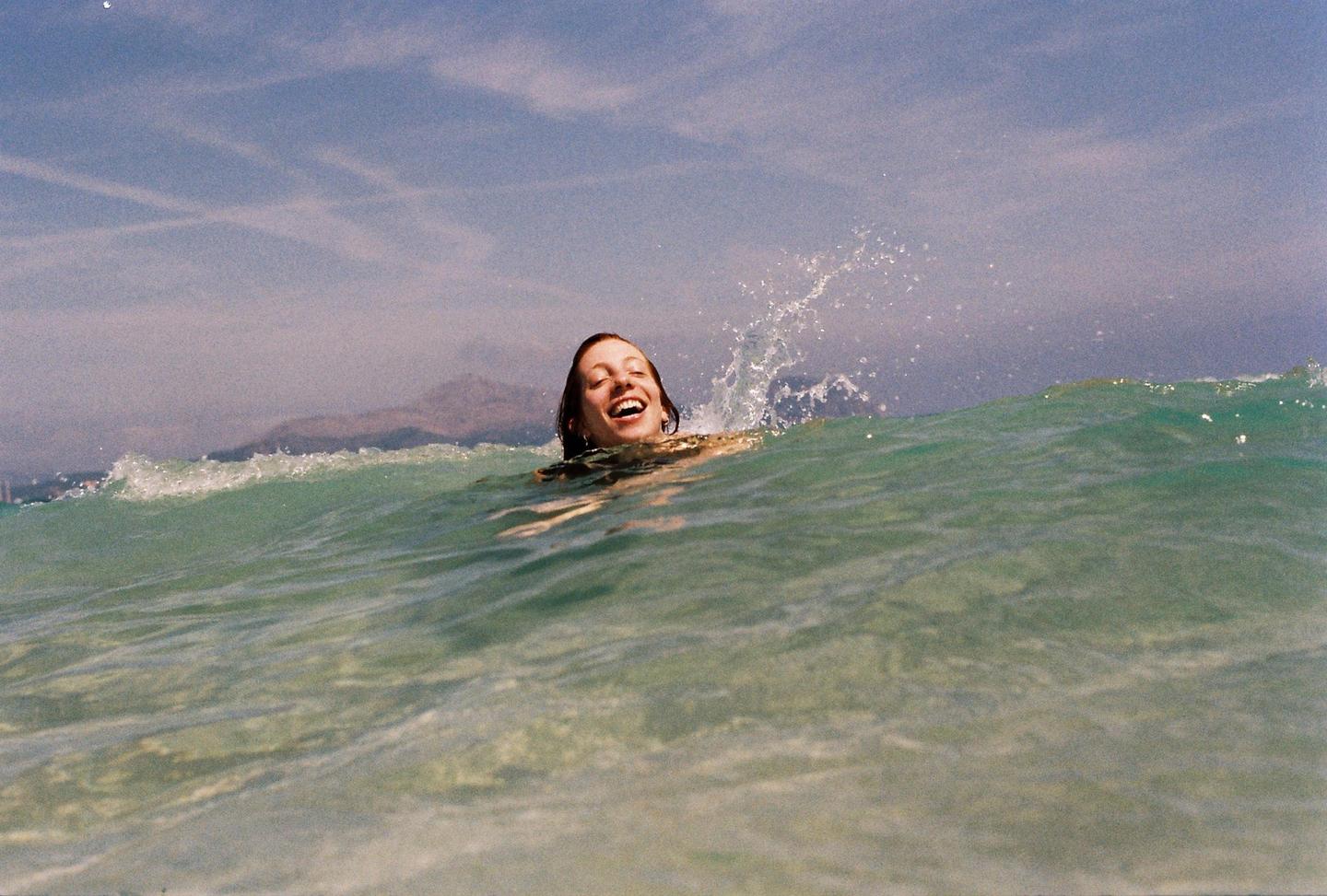 A woman with a joyful expression is in the ocean with water splashing around her.