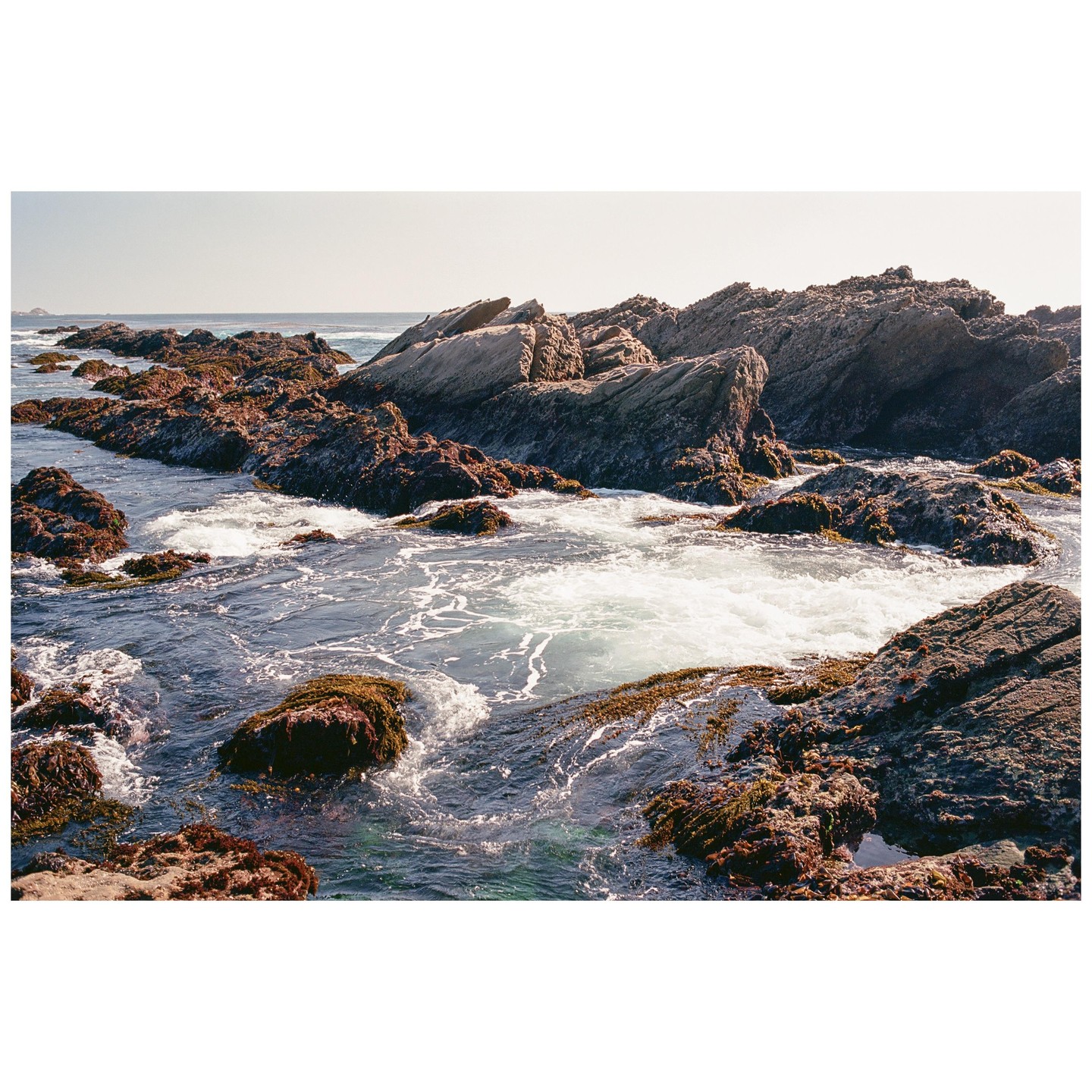 Waves crash against a rocky coastline covered in seaweed under a clear sky.