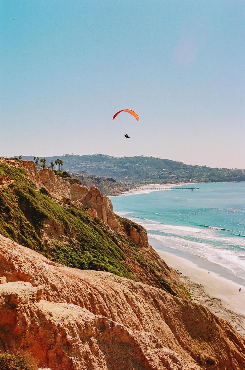 A paraglider soars above a cliff overlooking a beach and ocean.