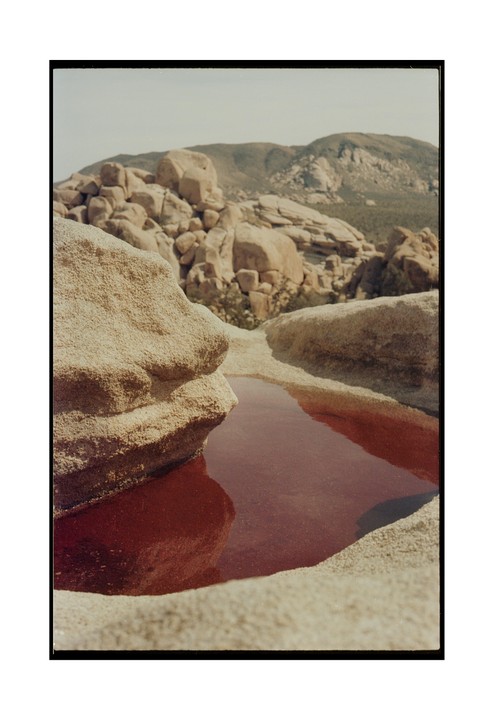 A pool of red water sits among desert rocks with mountains in the background.