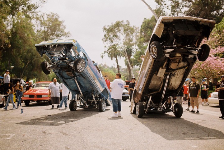 Two lowrider cars are on display with their front ends lifted high into the air, surrounded by people in a park.