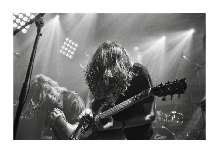 A man with long hair plays an electric guitar on a stage with another musician in the background.