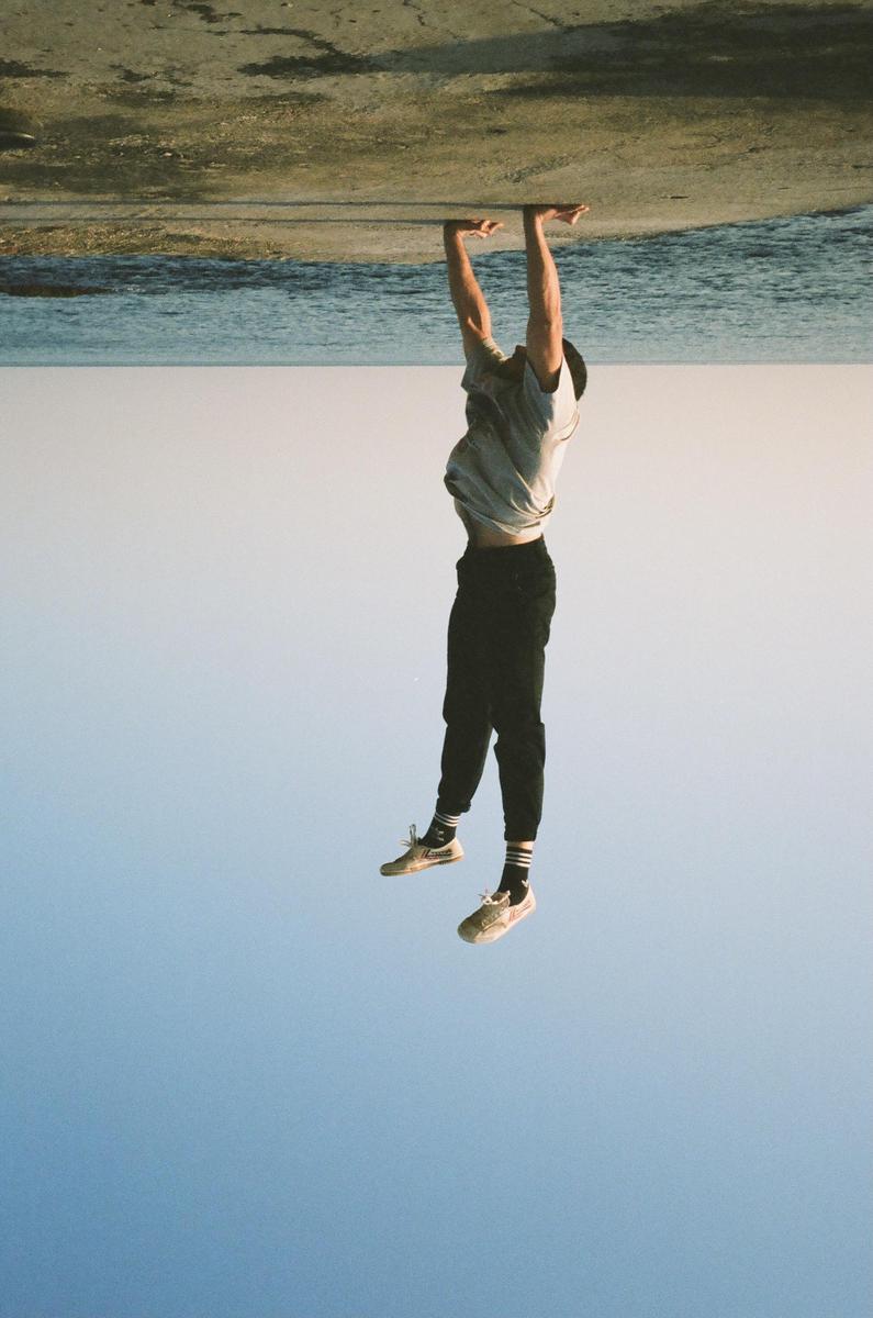 A man is doing a handstand on a concrete surface next to water, with the sky below him.