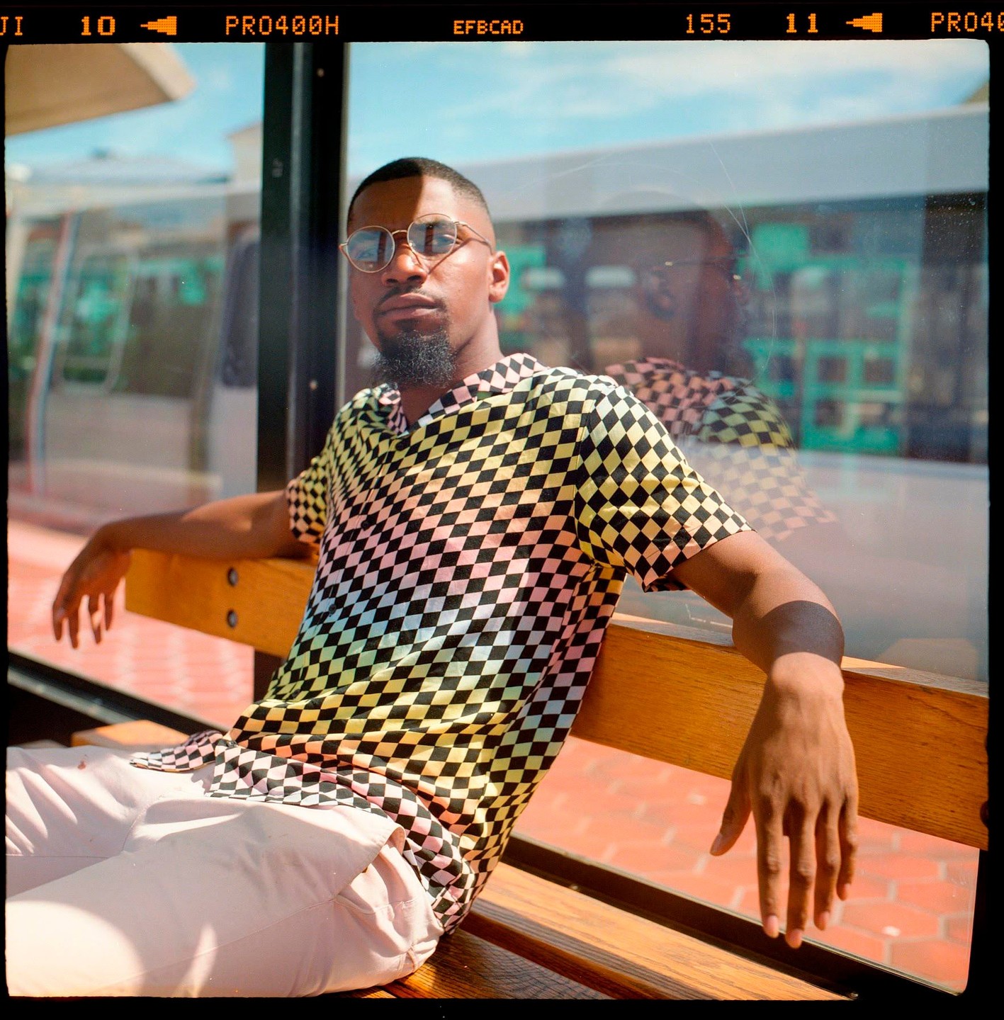 A man wearing a colorful checkered shirt and sunglasses sits on a wooden bench.