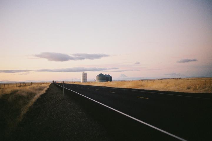 A long, empty road stretches into the distance with fields of dry grass on either side, leading to a cluster of silos and a distant mountain under a pale sky.