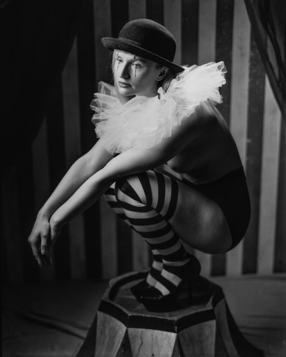 A woman in a bowler hat and striped stockings crouches on a circus prop.