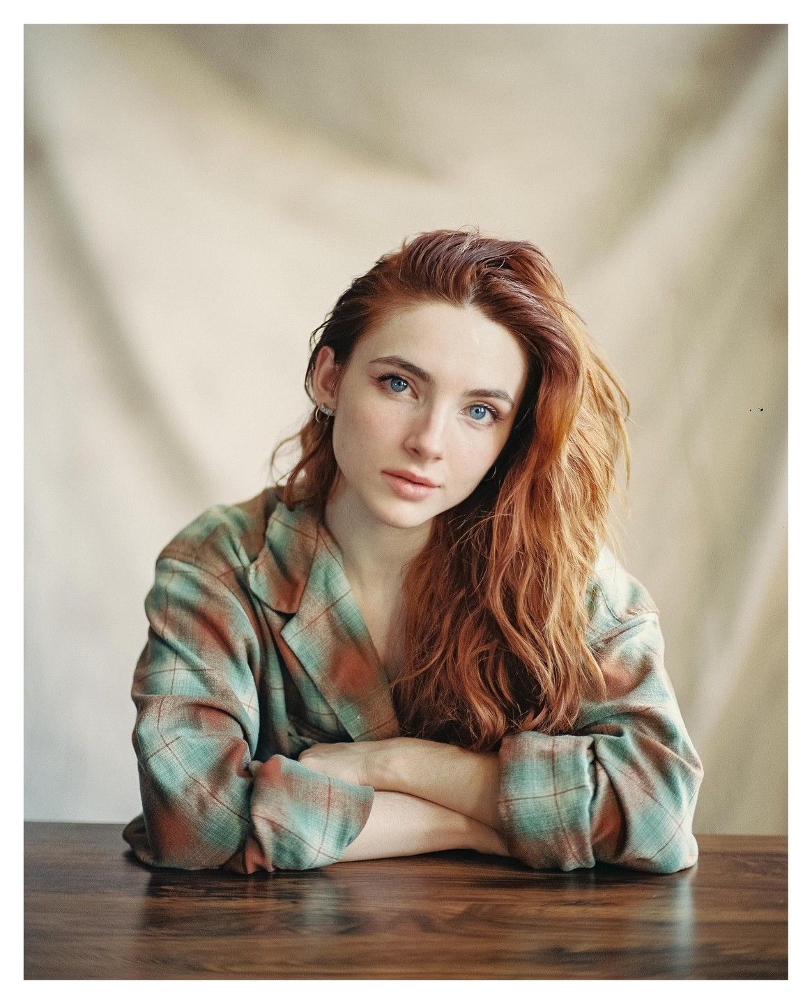A woman with long red hair and a plaid shirt sits at a wooden table with her arms crossed.