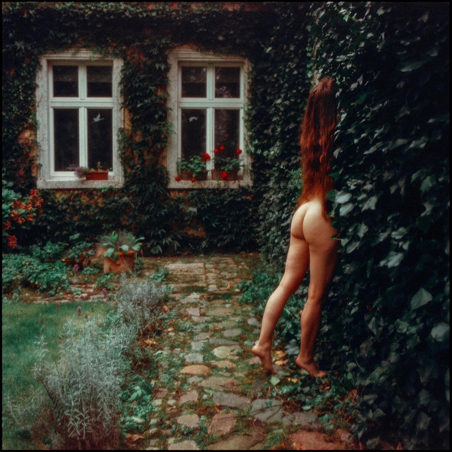 A nude woman with long red hair stands next to an ivy-covered wall with two windows, with a stone path and garden in the foreground.